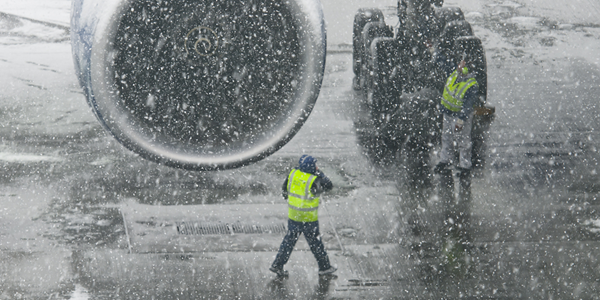 Two airport employees on a tarmac during a snow storm. They are wearing high visibility vests and standing near a giant aircraft.