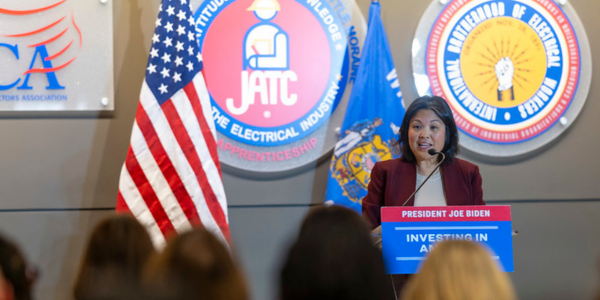 Acting Secretary Su speaks at a podium. On the stage behind her are an American flag and seals representing various trades. 