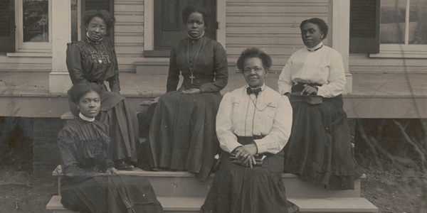 Sepia photo: Nannie Helen Burroughs and four other Black other women in Victorian dresses sit on the stairs to the front porch of the National Training School in Washington, D.C. Library of Congress photo.