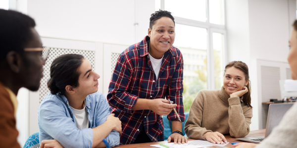 A diverse ethnic group of young students seated at a table and working with a smiling instructor in a plaid shirt.