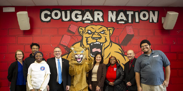 Acting Secretary Su and Secretary of Education Miguel Cardona pose for a photo with Job Corps staff and students in front of a mural reading âCougar Nation.â