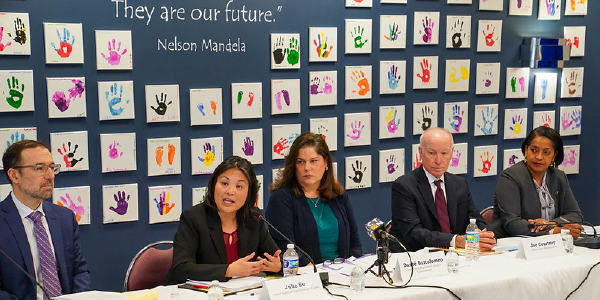 Acting Secretary Julie Su speaks in a roundtable, surrounded by colleagues, in front of a wall bearing colorful paint handprints of children.