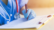 A medical professional in scrubs and gloves enters information on a clipboard. 