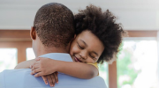 A child with curly hair hugging his dad around the neck.