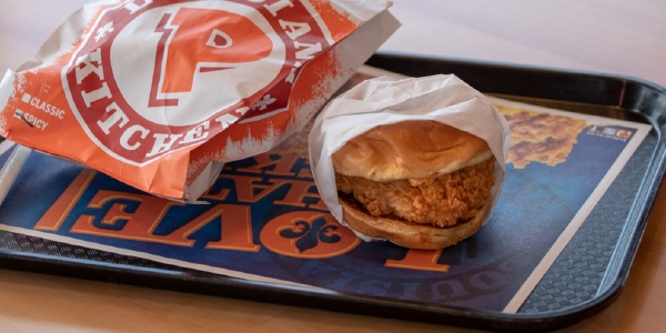 A fried chicken sandwich on a plastic tray next to a paper bag with the Popeyes logo. 