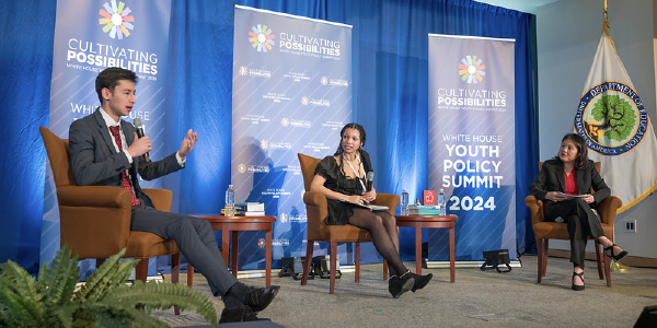 Acting Secretary Su joins a young man and a young woman on stage for a conversation. Banners behind them read: âCultivating Possibilities: Youth Policy Summit 2024â