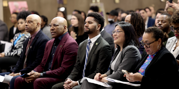 Acting Secretary Su smiles while listening to a speaker. In a full auditorium, she is seated in the front row alongside several Black guests, all listening attentively.