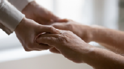 Two people, one older and seated, clasp hands in a supportive gesture.