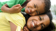 A Black mother wearing a yellow shirt laying in the grass with her son laying on her back wearing a green shirt as they smile for a photo.