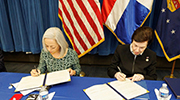 Two women sign official papers, seated in front of the flags of the United States, the Dominican Republic and the Labor Department.