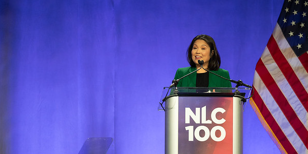 Acting Secretary Su smiles while standing at a podium with an NLC 100 logo on it. An American flag is on the stage behind her. 