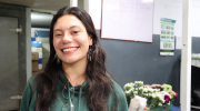 A young woman in a green sweatshirt smiles in a flower shop, in front of a tray of bouquets.