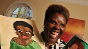 Lois Curtis, a Black woman with short hair, smiles broadly while holding up a self-portrait.