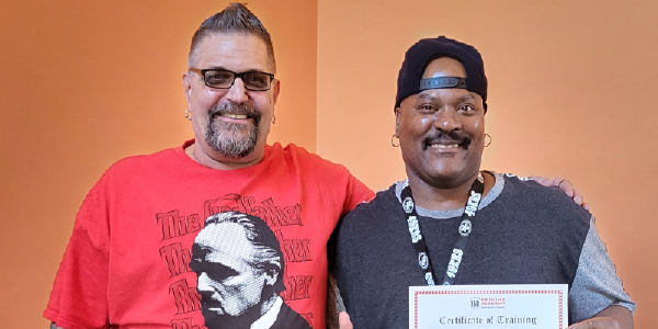 Les and Michael, two adult men, grin broadly while posing for a photo. Les has a hand on Michaelâs shoulder. Michael holds up a certificate of training. 