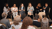 Six diverse women sit in chairs on a stage holding microphones for a panel discussion