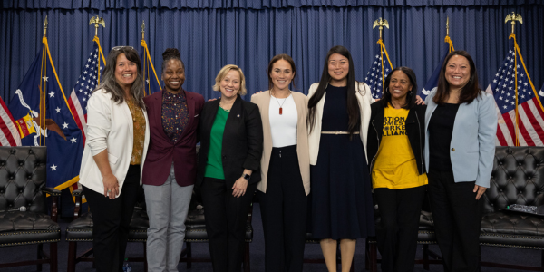 Seven diverse women stand together in front of a blue curtain, leather seats and various American flags while smiling for a photo. 