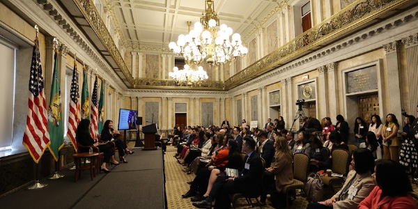 Acting Secretary Su is seated on a low stage in the White House for an armchair conversation. Chandeliers hang over a seated crowd.