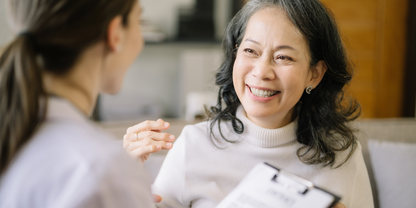 An Asian woman with dark, shoulder length hair and wearing a cream-colored turtleneck sweater smiling and looking at her doctor who is a woman with dark hair styled in a low ponytail while wearing a white coat and holding a clipboard.