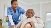 An older Black man seated on a couch and holding a cane while looking up towards a Black woman nurse in blue scrubs who is smiling and touching his shoulder.