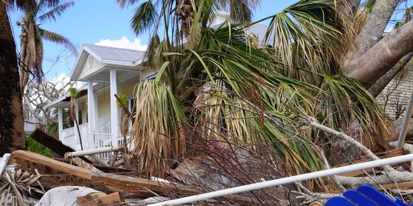 Damage from a recent storm showing a white house with debris and fallen trees around it, including scattered broken items and a blue chair in the foreground.