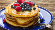 A stack of pancakes topped with fresh blueberries, raspberries, and a light drizzle of syrup, served on a blue plate with a fork on the side, placed on a wooden table.