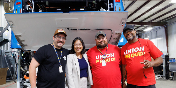 Acting Secretary Su stands with three men, two wearing âUnion Proudâ tee shirts, in front of an electric bus whose back panel is raised to show the inner workings. 