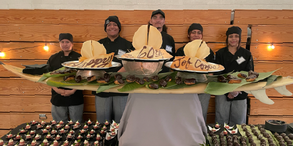Five Job Corps students pose behind a table carrying a celebratory platter of hors dâoeuvres theyâve prepared. The words Happy 60th Job Corps are written in icing on three desserts. 