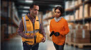 A man and woman speak inside a warehouse. Both are wearing orange for visibility. 