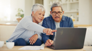 An elderly couple with grey hair seated use a laptop while reviewing financial documents.