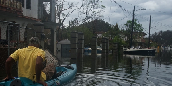 A Black man in a yellow shirt paddles a kayak through a flooded suburban street in Florida. Water covers  the road, and a boat floats in a yard. Photo by Eliud Echevarria, FEMA