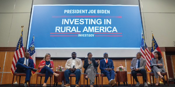 Acting Secretary of Labor Julie Su sits on a stage with six men and women in a panel discussion. Behind  them is a large sign reading President Joe Biden Investing in Rural America. Invest.gov