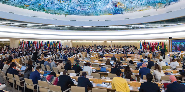 An ILO conference room in Switzerland shows dozens of people seated in concentric circles under a colorful ceiling. The walls are lined with flags from many nations. 