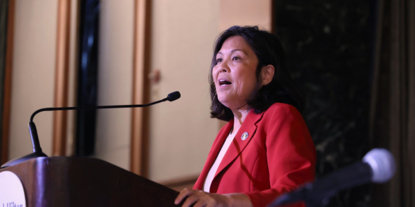 Acting Secretary Julie Su stands at a podium wearing a white shirt and red blazer as she addresses a room. 