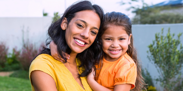 A mother with long, dark hair standing and holding her daughter while embracing each other and smiling for a photo. 