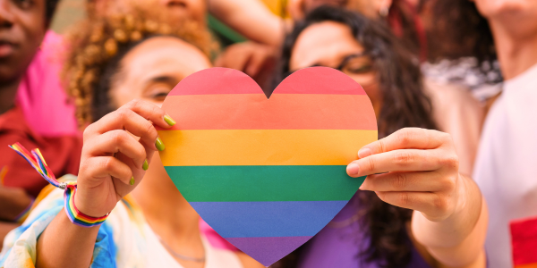 A diverse group of people gathered together with two women standing in front and holding a rainbow heart. 