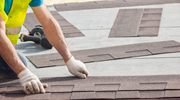 A roofer wearing work gloves places shingles on a roof. 