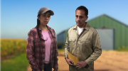 A man holding documents speaks with a woman farmworker near a barn. 
