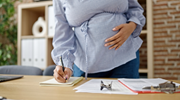 A pregnant woman at work writes in a notebook on a desk. 