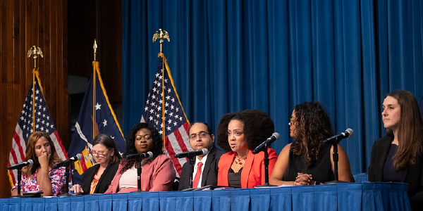 Seven professionally dressed women and men sit on a panel behind a table. American and Labor Department flags are visible on the stage behind them.