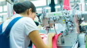 A person using an industrial sewing machine to stitch a bright pink fabric in a manufacturing setting. The focus is on their hands guiding the material through the machine.