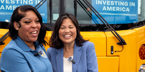 Acting Secretary Su smiles with a woman in a blue shirt. They stand in front of a yellow school bus with signs reading “Investing in America” in the windows behind them. 