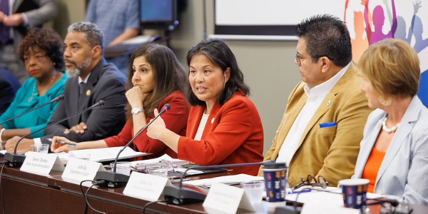 Acting Secretary Su and Domestic Policy Advisor Neera Tanden, both wearing red suits, participate in a roundtable discussion at a table with microphones. Several elected officials and community leaders are seated around them.
