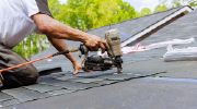 A roofer kneels on a roof, installing new shingles with a nail gun.