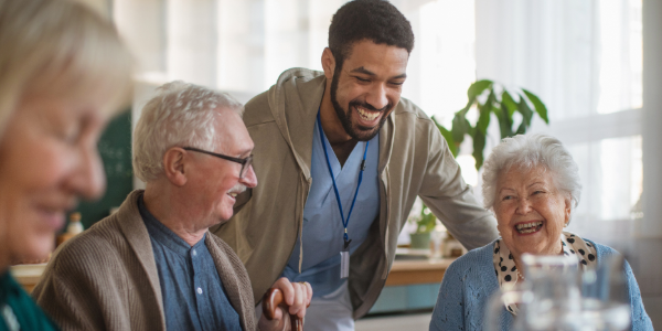 A caregiver laughs joyfully with three elderly individuals around a table in a bright room.