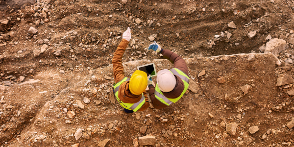 Two individuals wearing hard hats, reflective vests and protective gear stand in a quarry and point into the distance while using a tablet.