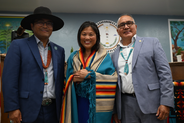 Acting Secretary Su stands between two Navajo men. The men are wearing beaded necklaces and Su has a colorful striped blanket draped around her shoulders. All three smile for the camera.