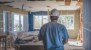 Construction worker in a hard hat inspecting a room under renovation, with exposed beams, hanging electrical wires, and construction materials scattered around.