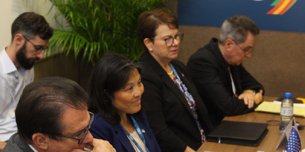 Acting Secretary Su listens to an unpictured speaker, seated at a conference table with four men and women in professional clothing.