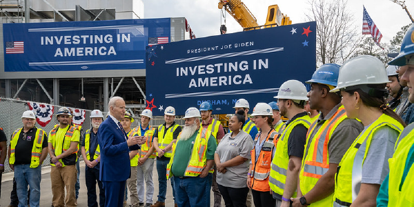 President Joe Biden addresses a dozen workers in high-visibility jackets and hard hats in front of construction equipment and signs reading âInvesting in America.â 