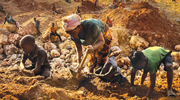 A woman and two children work in an above-ground mine, with other workers in the background. She uses a shovel, and they use their hands to dig through dirt and rocks.